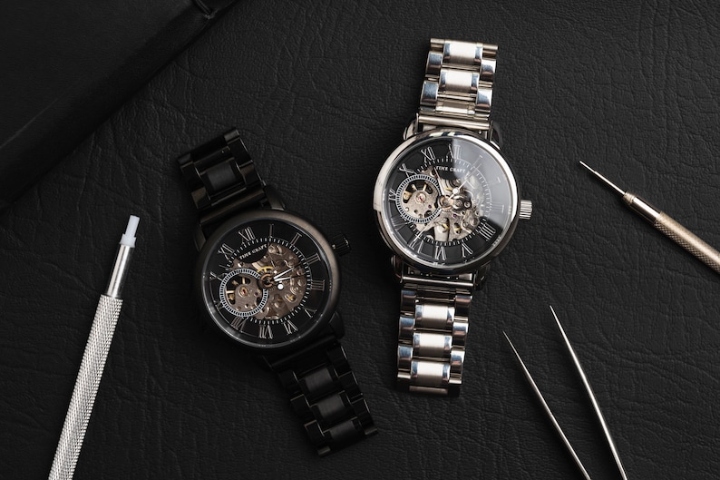 May include: Two skeleton watches with black faces and Roman numeral hour markers. The watch on the left has a black metal band and the watch on the right has a silver metal band. Both watches are on a black leather surface.