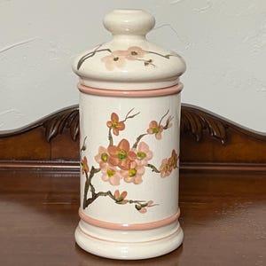 May include: A white ceramic jar with a pink lid and floral design. The jar features a pink and green floral pattern with a pink rim around the top and bottom.