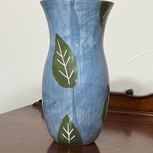 May include: A ceramic vase with a flared rim and a tapered body, featuring a light blue base with a brushstroke texture and dark green leaf motifs painted on the surface, each leaf detailed with thin white lines representing the veins.