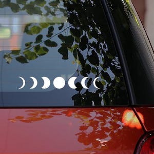 Op de afbeelding: Een witte vinyl sticker met de maanfasen is bevestigd op de achterruit van een rode auto. De fasen lopen van de halve maan naar de volle maan en terug. De ruit van de auto weerspiegelt een boom en de lucht.