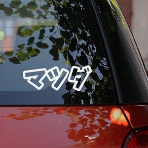 May include: A white vinyl sticker with Japanese characters is applied to the rear window of a red car. The sticker is outlined in white. The background shows a blurred view of trees and other cars. The car's taillight is visible.