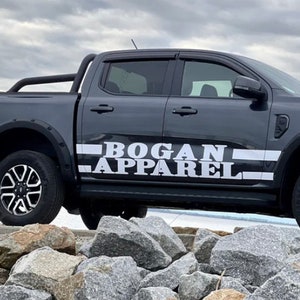 May include: A dark gray pickup truck with a black roll bar and a white "BOGAN APPAREL" decal on the side. The truck is parked on a rocky surface.