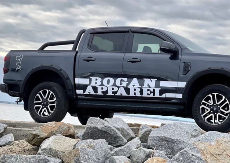 May include: A dark gray pickup truck with a black roll bar and a white "BOGAN APPAREL" decal on the side. The truck is parked on a rocky surface.