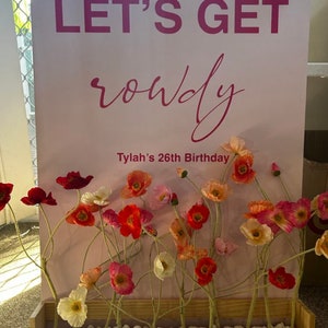 May include: A pink sign with the text "Let's Get Rowdy" and "Tylah's 26th Birthday" surrounded by a floral arrangement of pink, red, and white poppy flowers in a wooden box.