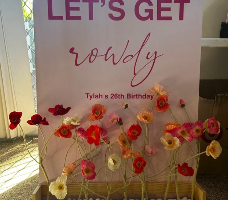 May include: A pink sign with the text "Let's Get Rowdy" and "Tylah's 26th Birthday" surrounded by a floral arrangement of pink, red, and white poppy flowers in a wooden box.