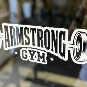 May include: White vinyl sticker with a black and white graphic of a barbell with weights on each end. The text "ARMSTRONG GYM" is written in a bold, black font above the barbell.
