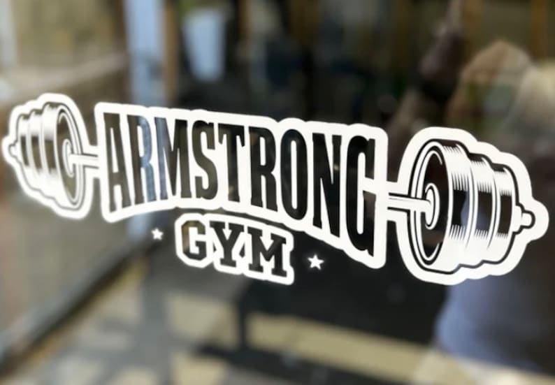 May include: White vinyl sticker with a black and white graphic of a barbell with weights on each end. The text "ARMSTRONG GYM" is written in a bold, black font above the barbell.