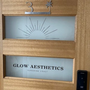 May include: A wooden door with a frosted glass panel featuring a stylized sunburst design. The door has a silver number "3" and a palm tree cutout above the glass panel. The bottom panel features the text "GLOW AESTHETICS SUNSHINE COAST".