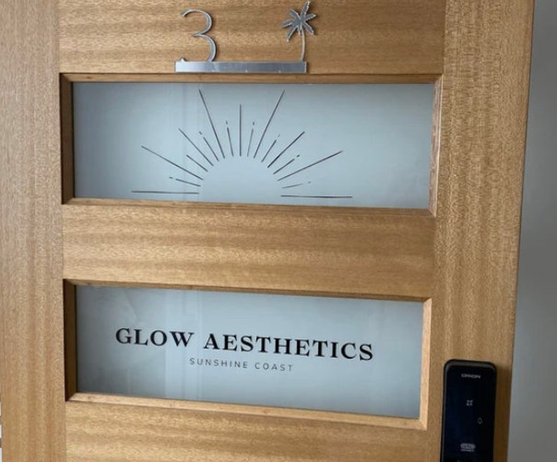 May include: A wooden door with a frosted glass panel featuring a stylized sunburst design. The door has a silver number "3" and a palm tree cutout above the glass panel. The bottom panel features the text "GLOW AESTHETICS SUNSHINE COAST".