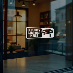 Op de afbeelding: Een rechthoekige witte raamsticker met een zwarte rand en zwarte tekst die SECURITY CAMERAS IN USE leest, vergezeld van een zwart grafisch icoon van een beveiligingscamera rechts van de tekst op een witte achtergrond.