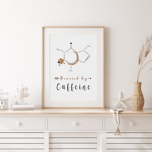Puede incluir: Impresión enmarcada que presenta un diagrama de la molécula de cafeína con un diseño de mancha de café. El texto "Powered by Caffeine" está debajo. La obra de arte está en un marco de madera clara.