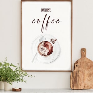 May include: A framed print with the text "But first, coffee" in brown lettering. A watercolor illustration of a cup of coffee with coffee beans on a white saucer is below the text.