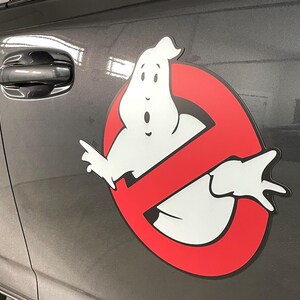 May include: A Ghostbusters logo decal applied to the side of a dark gray vehicle. The design features a white cartoon ghost centered within a red circle with a diagonal slash, all outlined in black, creating a high-contrast graphic display.