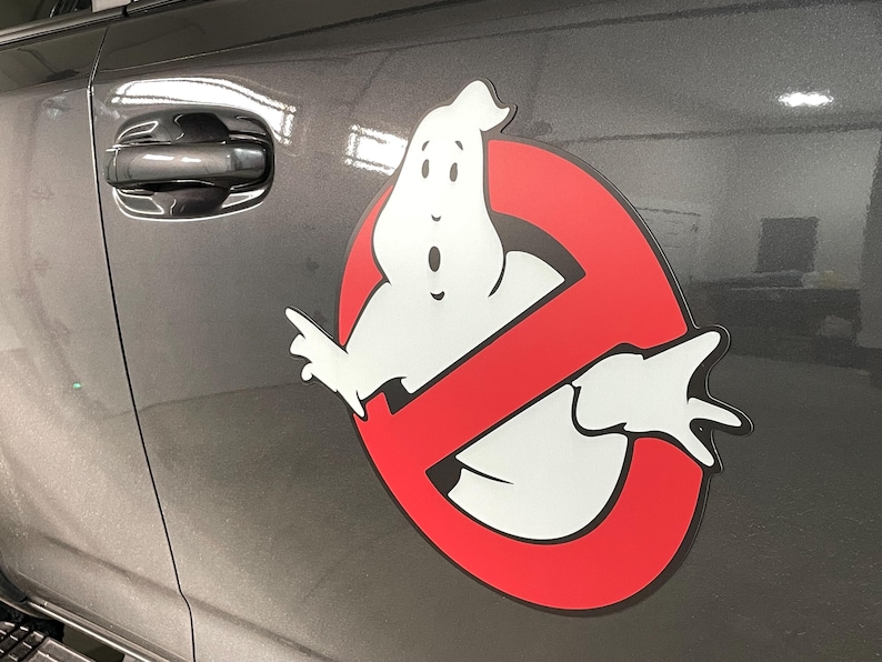 May include: A Ghostbusters logo decal applied to the side of a dark gray vehicle. The design features a white cartoon ghost centered within a red circle with a diagonal slash, all outlined in black, creating a high-contrast graphic display.