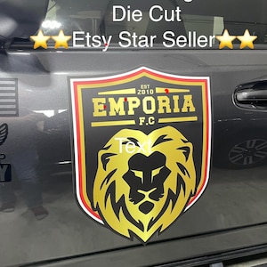May include: A die-cut business magnet featuring a shield-shaped emblem with a gold lion head graphic. The text reads "EST 2010", "EMPORIA", and "F.C." in gold lettering against a black background, bordered by thin red, white, and gold lines.