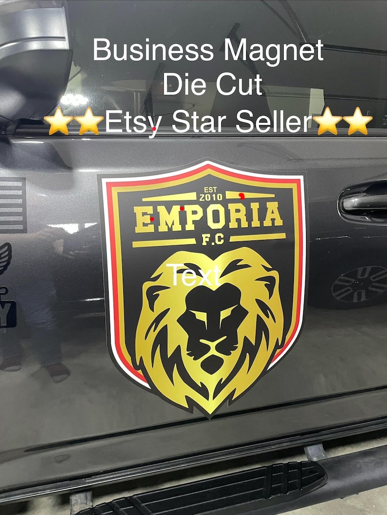May include: A die-cut business magnet featuring a shield-shaped emblem with a gold lion head graphic. The text reads "EST 2010", "EMPORIA", and "F.C." in gold lettering against a black background, bordered by thin red, white, and gold lines.