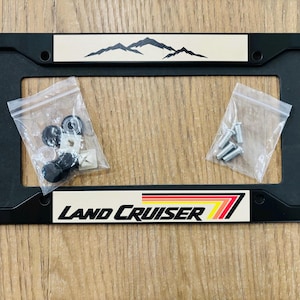 May include: Black license plate frame with the words "LAND CRUISER" and a mountain graphic. Includes hardware in small plastic bags. The frame is rectangular and has a black finish.