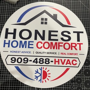 May include: A circular white sign featuring a black house icon, the text HONEST HOME COMFORT, and the slogan HONEST ADVICE. | QUALITY SERVICE. | REAL COMFORT. It includes the phone number 909-488-HVAC and a blue and red snowflake and sun graphic.