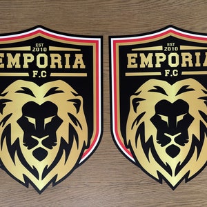 May include: Two identical shield-shaped emblems featuring a gold lion head centered on a black background. Above the lion, the text reads EMPORIA F.C. with EST 2010 above it. The shield has a thick black border with thin white and red accent lines.