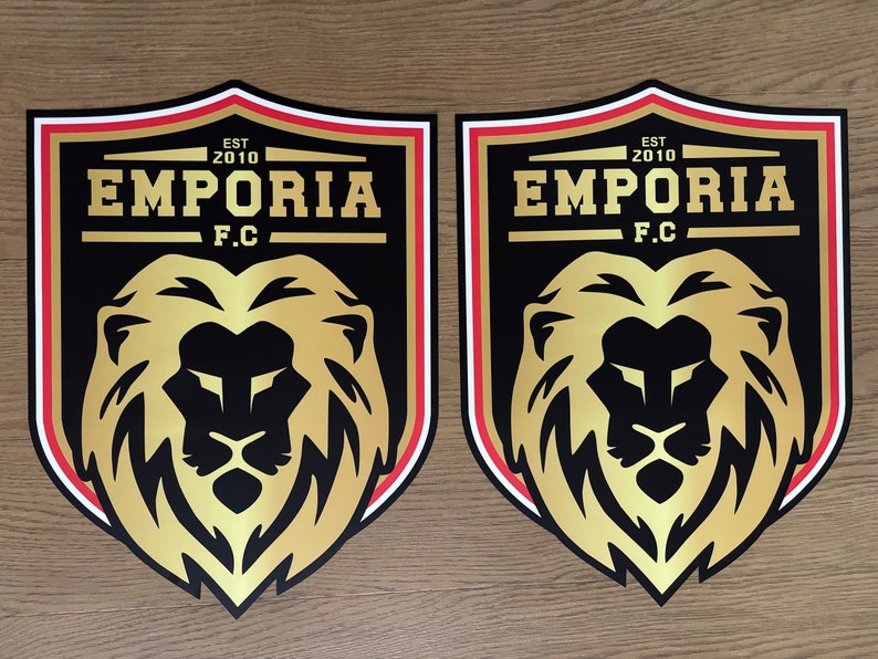 May include: Two identical shield-shaped emblems featuring a gold lion head centered on a black background. Above the lion, the text reads EMPORIA F.C. with EST 2010 above it. The shield has a thick black border with thin white and red accent lines.