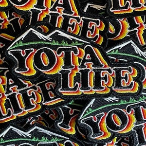 May include: Black embroidered patch with the words "YOTA LIFE" in a stylized font. The letters are outlined in white with a gradient of yellow, orange, and red. A green mountain range design is above the text.