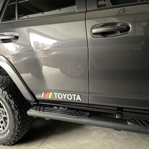 May include: A gray Toyota SUV with black side steps and a red, yellow, and black "TOYOTA" decal on the side.