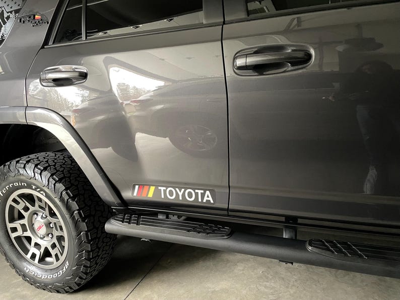 May include: A gray Toyota SUV with black side steps and a red, yellow, and black "TOYOTA" decal on the side.