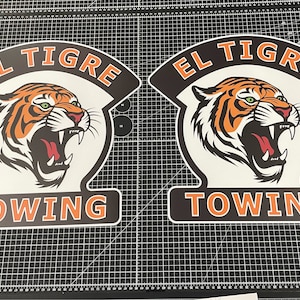 May include: Two identical custom decals featuring a snarling tiger head graphic in orange, black, and white. The text EL TIGRE is arched above the tiger, and the word TOWING is printed in a bold orange font at the bottom of each shield-shaped sign.