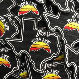 May include: Close-up of several black fabric patches shaped like the state of Texas. Each patch features a stylized sunset with red, orange, and yellow stripes and the text "YOTA LIFE" in white.