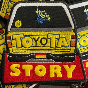 May include: A yellow and black embroidered patch with the word "TOYOTA" and a cartoon alien character. The patch also has a red and yellow embroidered banner with the word "STORY" on it.