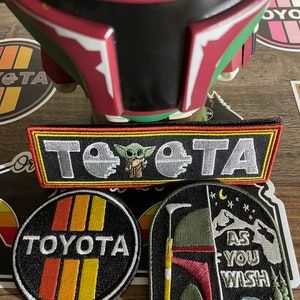 May include: Three embroidered patches featuring Star Wars characters. The first patch is black with yellow and orange stripes and the word "TOYOTA" in white. The second patch is black with a white outline and features a green alien character with a lightsaber and the text "TOYOTA". The third patch is green and brown with a white outline and features a helmet with the text "AS YOU WISH".