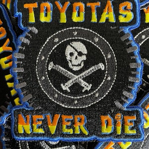 May include: A black embroidered patch with a skull and crossbones design. The patch has a white skull with a black eyepatch and crossed bones. The text "TOYOTAS NEVER DIE" is embroidered in yellow and orange on the patch.