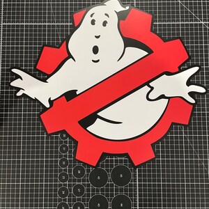 May include: A flat, die-cut sign featuring the iconic white ghost character with black outlines, positioned in front of a red gear-shaped symbol with a diagonal red slash. The design is centered on a black cutting mat with white grid lines.