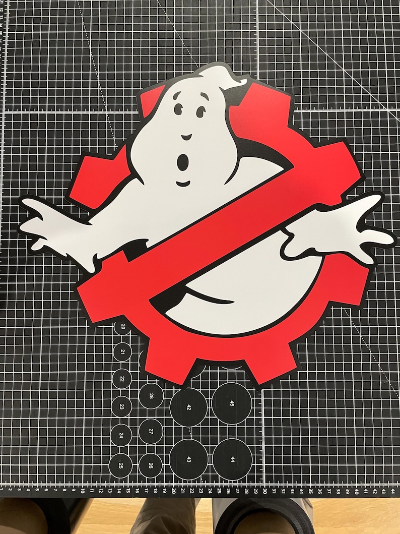 May include: A flat, die-cut sign featuring the iconic white ghost character with black outlines, positioned in front of a red gear-shaped symbol with a diagonal red slash. The design is centered on a black cutting mat with white grid lines.