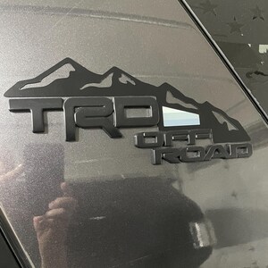 May include: A matte black TRD Off Road emblem featuring a mountain range silhouette above the lettering. The emblem includes a small rectangular accent with horizontal stripes in white, light gray, and dark gray, applied to a dark gray vehicle surface.