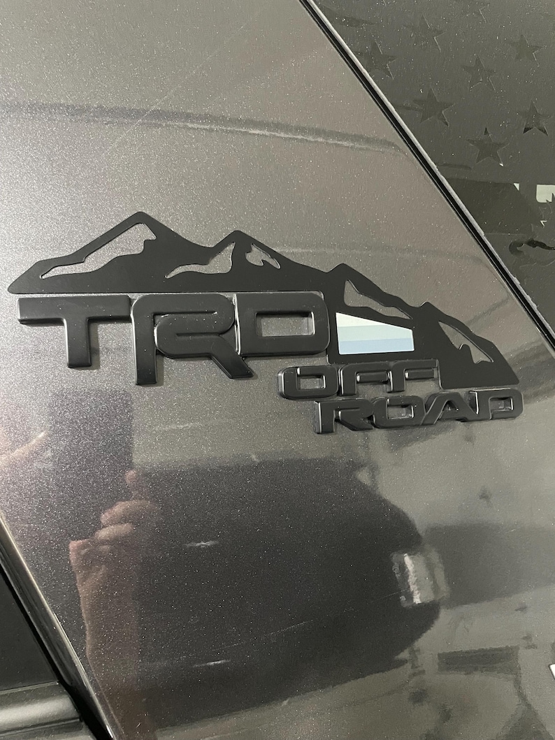 May include: A matte black TRD Off Road emblem featuring a mountain range silhouette above the lettering. The emblem includes a small rectangular accent with horizontal stripes in white, light gray, and dark gray, applied to a dark gray vehicle surface.