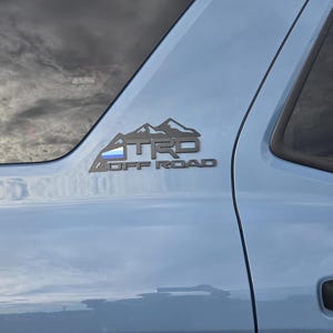 May include: A light blue vehicle door with a black TRD Off Road emblem. The emblem features a mountain range design above the text. The vehicle's door handle and window frame are visible.