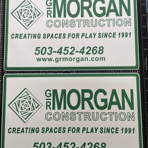 May include: Two rectangular white signs with dark green borders and text. Each sign features a geometric logo, the text GR MORGAN CONSTRUCTION, the slogan CREATING SPACES FOR PLAY SINCE 1991, the phone number 503-452-4268, and the website www.grmorgan.com.