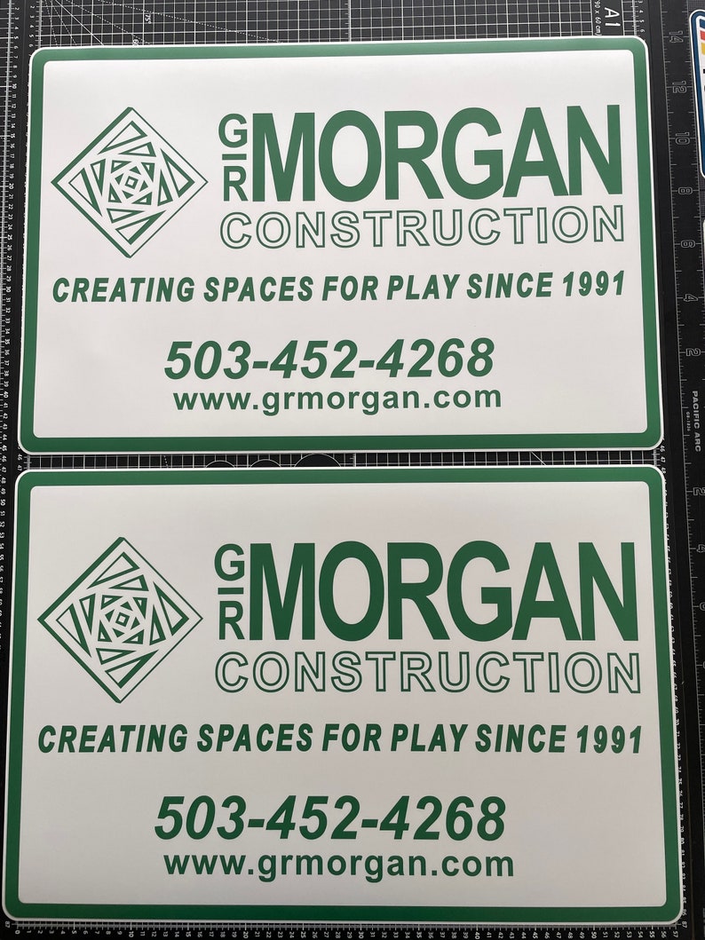 May include: Two rectangular white signs with dark green borders and text. Each sign features a geometric logo, the text GR MORGAN CONSTRUCTION, the slogan CREATING SPACES FOR PLAY SINCE 1991, the phone number 503-452-4268, and the website www.grmorgan.com.