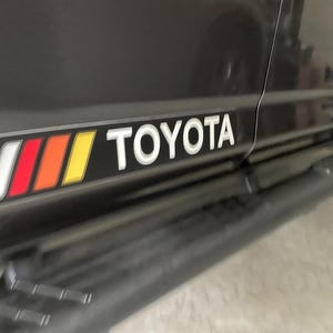 May include: A black Toyota truck with a red, orange, and yellow stripe and the word "TOYOTA" in white lettering on the side.