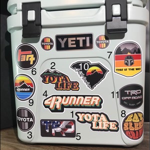 May include: A light blue Yeti cooler with various stickers on it. The stickers include a South Carolina outline, a Toyota logo, a 4Runner logo, a Yota Life logo, a TRD Off Road logo, and a Eat Sleep Crawl logo.