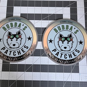 May include: Two circular stickers featuring a central illustration of a dog wearing goggles. The text BUBBA'S PATCHES is printed in black and green. The design includes two black stars, a forest silhouette at the bottom, and the URL WWW.BUBBASPATCHES.COM.