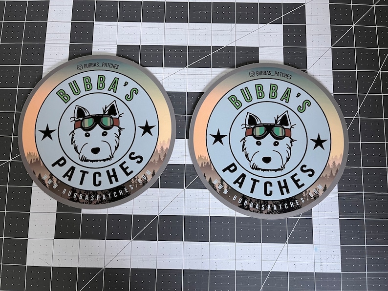 May include: Two circular stickers featuring a central illustration of a dog wearing goggles. The text BUBBA'S PATCHES is printed in black and green. The design includes two black stars, a forest silhouette at the bottom, and the URL WWW.BUBBASPATCHES.COM.