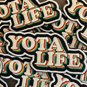 May include: A close-up of several embroidered patches with the text "YOTA LIFE" in a rainbow color scheme. The patches are black with white lettering and a white outline.