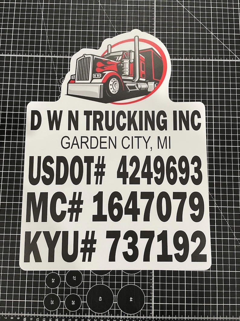 May include: A white rectangular sign with rounded top corners featuring a red and black semi-truck graphic. The text reads D W N TRUCKING INC, GARDEN CITY, MI, USDOT# 4249693, MC# 1647079, and KYU# 737192 in bold black lettering on a white background.