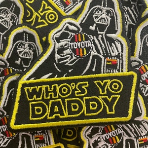 May include: A black and white embroidered patch featuring Darth Vader holding a Toyota logo. The patch has a yellow border and the text "Who's Yo Daddy" in yellow lettering.