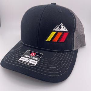 May include: Black trucker hat with a gray mesh back and a curved brim. The hat features an embroidered mountain design with yellow, orange, and red stripes. The Richardson brand label is visible.