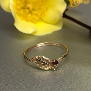 May include: A thin gold ring featuring a textured leaf motif on one side and a small, round, deep red gemstone set at the base of the leaf. The band is smooth and circular, showcasing a delicate design with a metallic finish and a dark red stone.