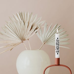 K&ouml;nnte beinhalten: Eine wei&szlig;e Kerze mit dem Text "&hearts; GL&Uuml;CKSUCH &hearts;" in schwarzen Lettern steht in einem terracottafarbenen Kerzenhalter. Der Kerzenhalter hat eine einzigartige ovale Form. Eine Vase mit getrockneten Palmbl&auml;ttern steht im Hintergrund.