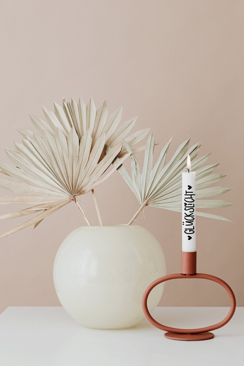 K&ouml;nnte beinhalten: Eine wei&szlig;e Kerze mit dem Text "&hearts; GL&Uuml;CKSUCH &hearts;" in schwarzen Lettern steht in einem terracottafarbenen Kerzenhalter. Der Kerzenhalter hat eine einzigartige ovale Form. Eine Vase mit getrockneten Palmbl&auml;ttern steht im Hintergrund.